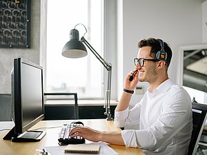 Ein Mann im weißen Hemd mit Headset sitzt an seinem Arbeitsplatz vor einem Bildschirm