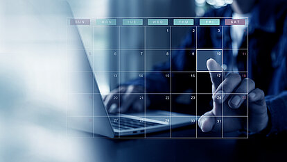 Eine Person sitzt vor einem Laptop und zeigt mit dem Finger auf einen digital dargestellten Kalender