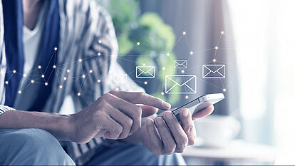 Eine Person tippt auf ihr Handy, über dem mehrere digital dargestellte Briefumschläge mit einem Verbindungspfad erscheinen